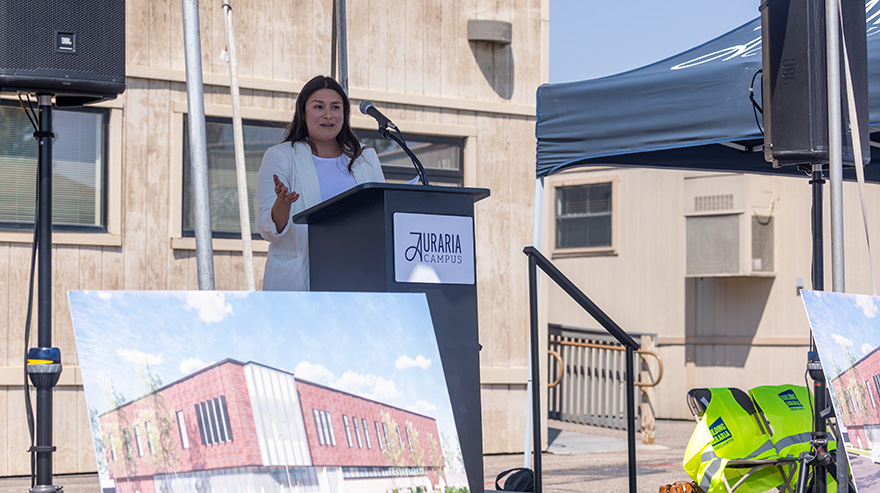 A speaker stands at a podium labeled "Auraria Campus" during an outdoor event with construction renderings displayed nearby.