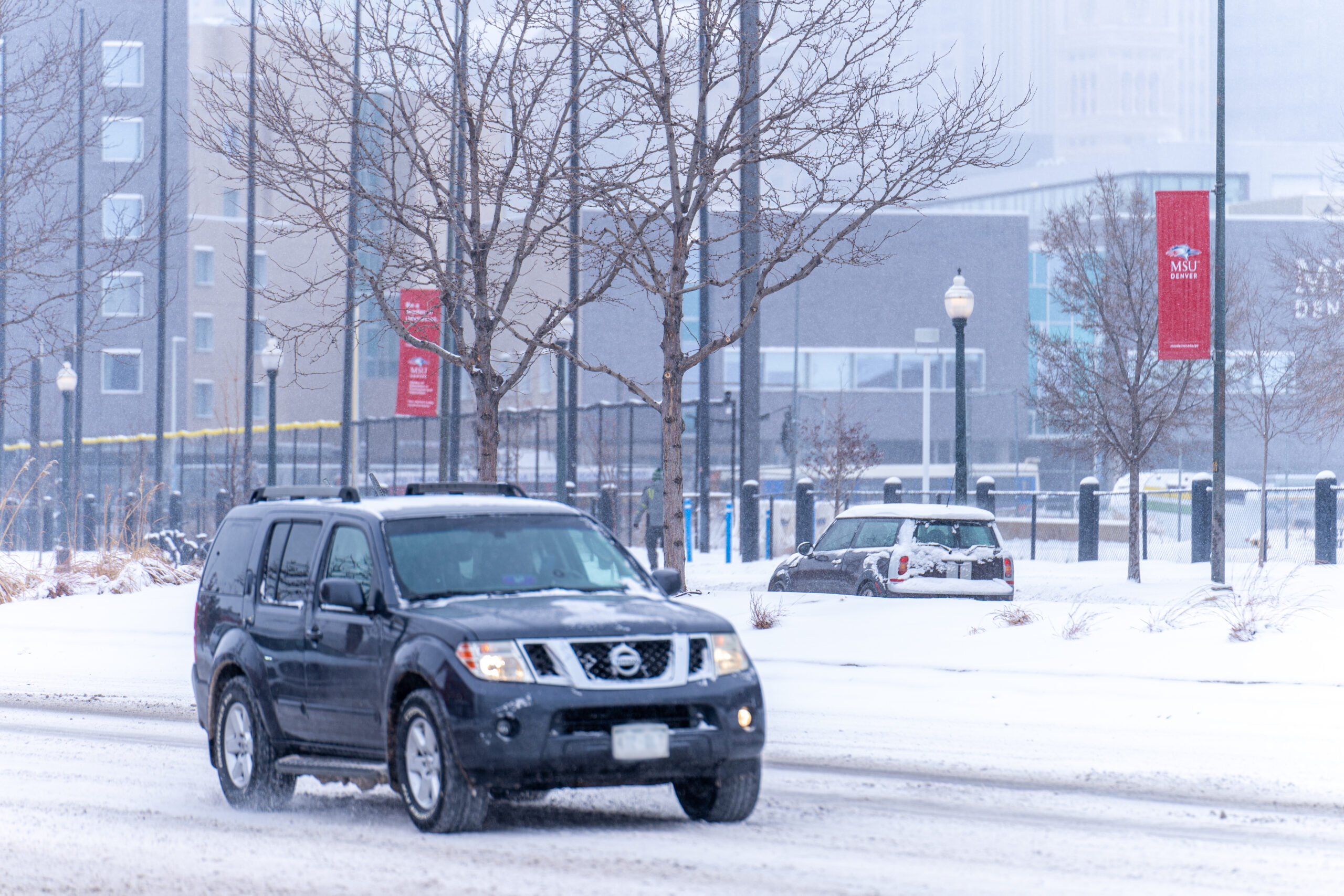 A vehicle drives down a snow-covered street on the MSU Denver campus during a winter storm, with campus buildings and red banners visible in the background.