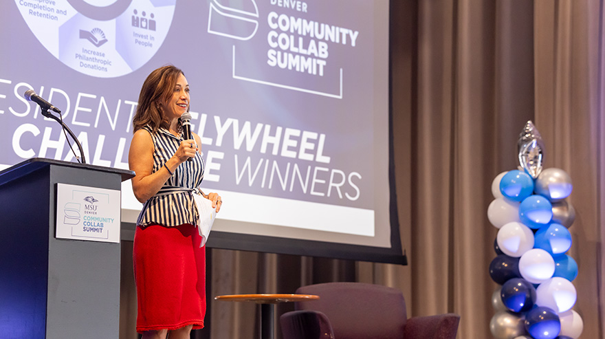 Dr. Janine Davidson speaks on stage at the Community Collab Summit beside a podium and large event screen.