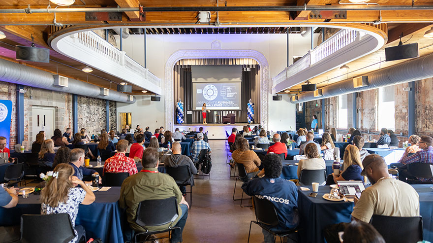 Summit attendees seated at tables watch Dr. Janine Davidson speak on stage.