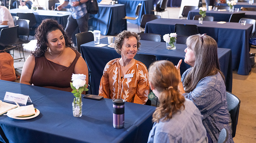 Four attendees in conversation at a table with blue tablecloths and flower vases.
