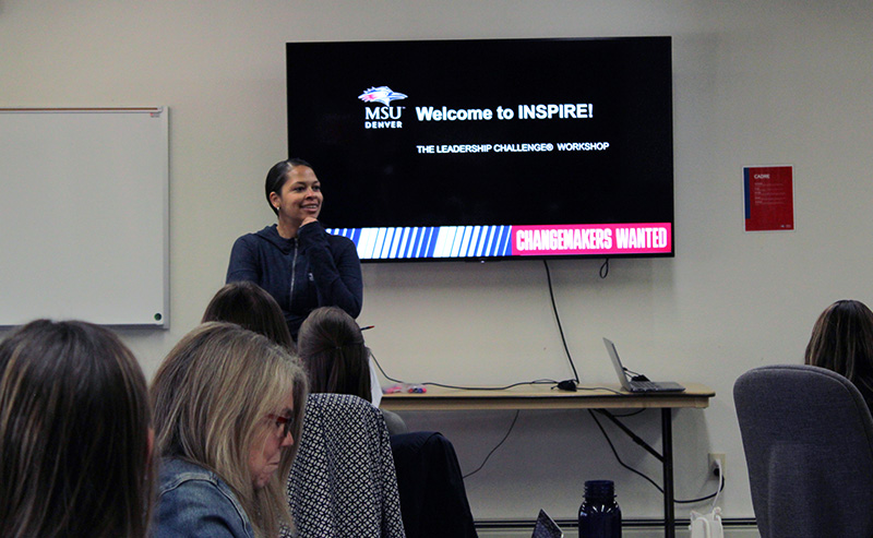 Imani Morning speaks at the front of a classroom during an MSU Denver leadership workshop. A screen behind her reads ‘Welcome to INSPIRE! The Leadership Challenge Workshop. Changemakers Wanted.’ People sit listening in the foreground.