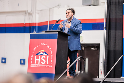 Michael Benitez stands at a lecture with HSI banner on the front, facing an audience.