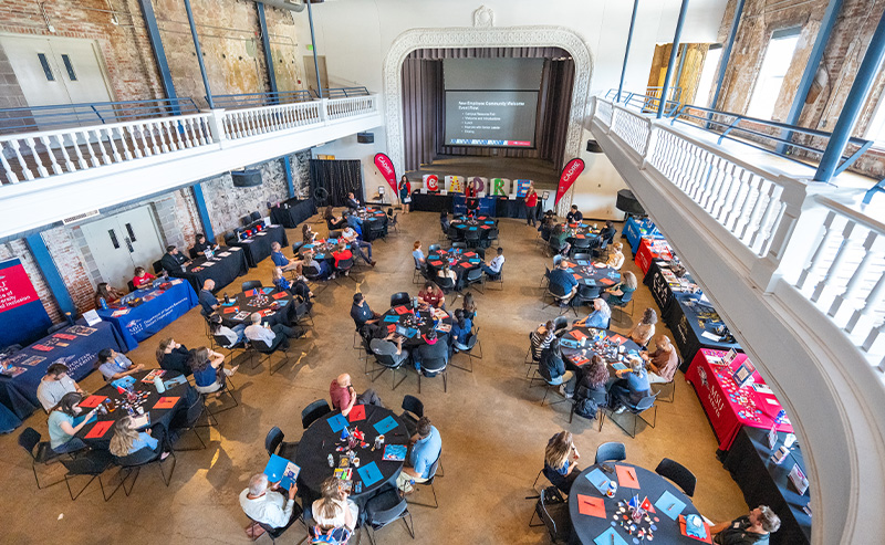 A wide view of MSU Denver’s New Employee Orientation shows attendees seated at round tables in a large hall, with a presentation screen and resource tables in the background.