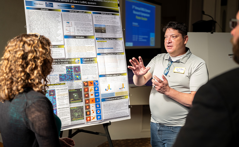 A faculty member presents research findings on a large, colorful poster board while engaging with two attendees at a symposium.