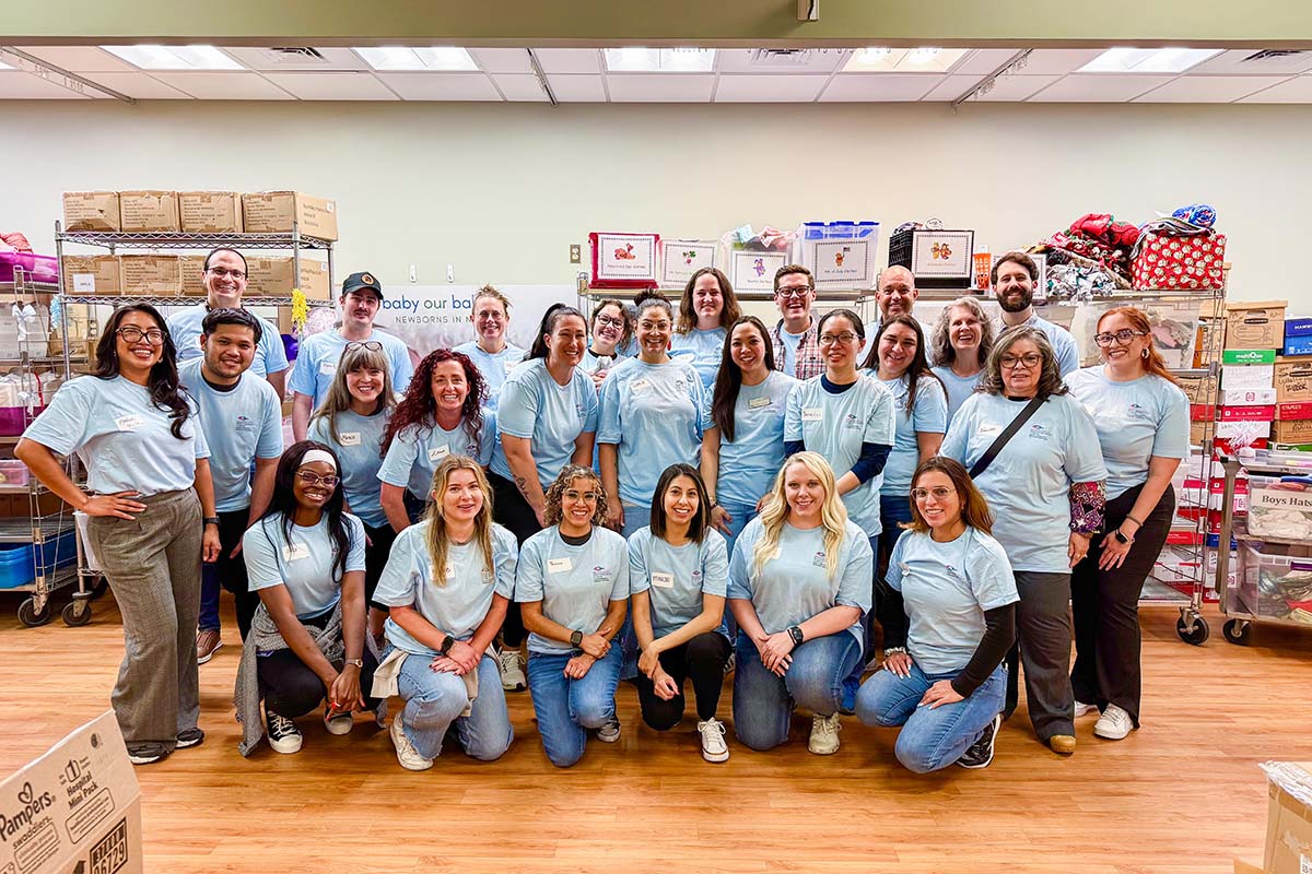 A large group of volunteers in blue shirts pose in a room filled with shelves and boxes of supplies.