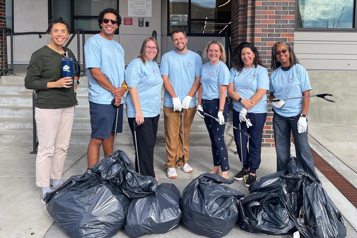 Seven volunteers in blue shirts pose with black trash bags on a sidewalk outside a brick building.