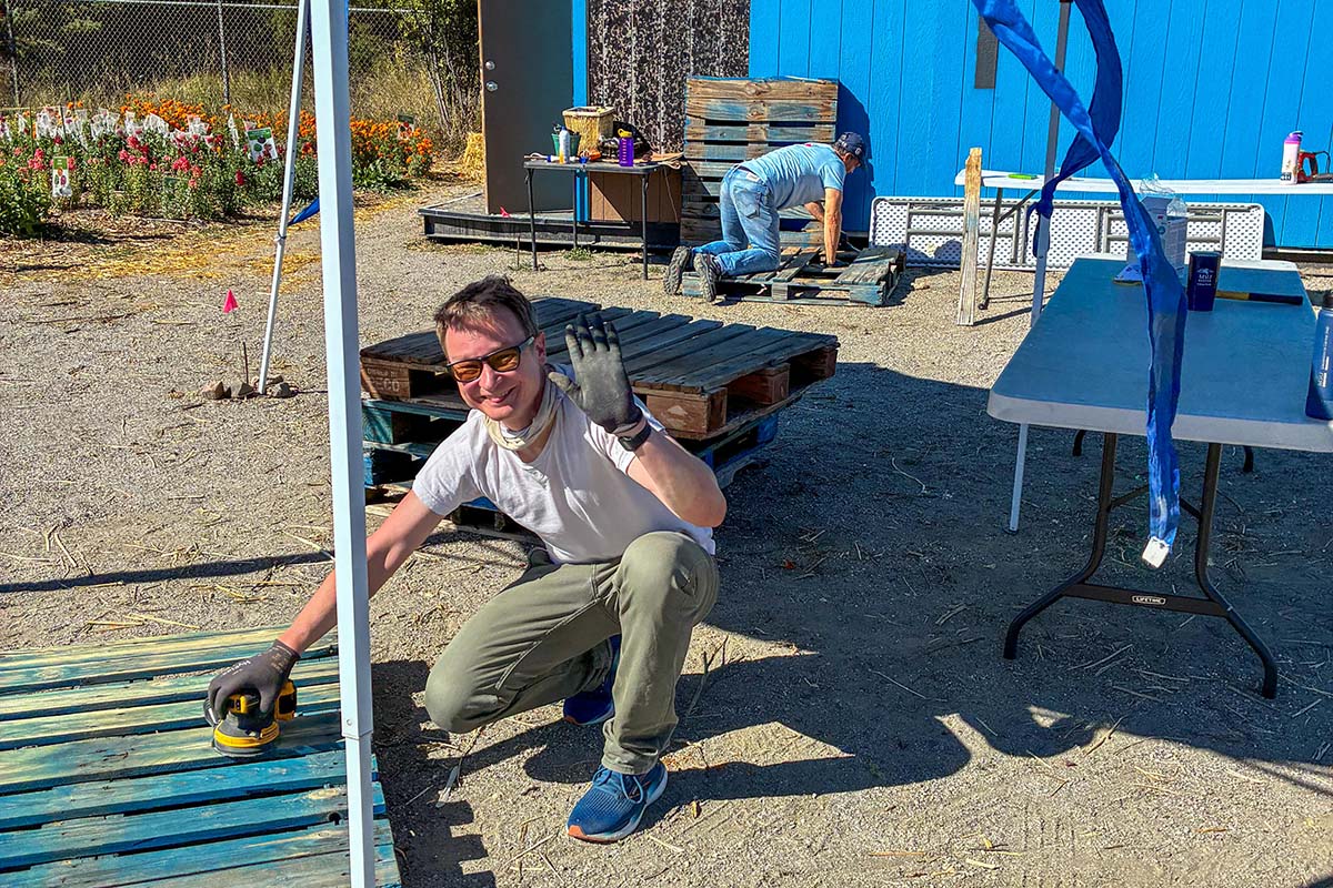 A volunteer wearing sunglasses kneels and waves while sanding a wooden pallet outside a blue building.