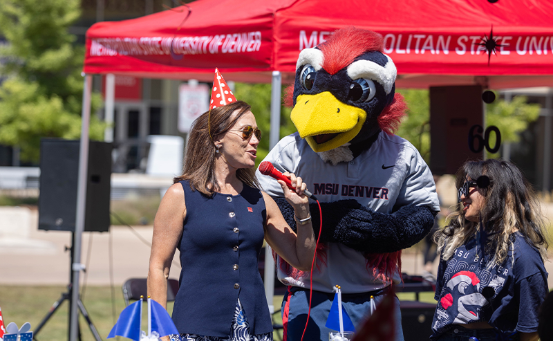 MSU Denver President Janine Davidson, wearing a party hat, speaks into a microphone alongside Rowdy the Roadrunner mascot during the university’s 60th birthday celebration at Welcome Week.