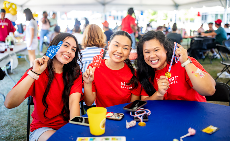 Three MSU Denver students in red shirts smile and hold up mini pennants while sitting at a blue table during Welcome Week’s Throwdown Thursday event