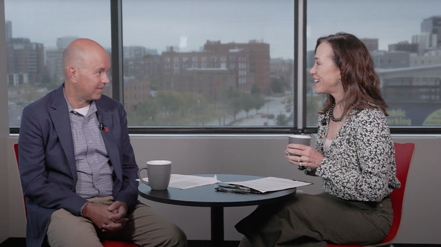 Two people seated at a table in front of a large window engage in conversation during an on-camera interview.