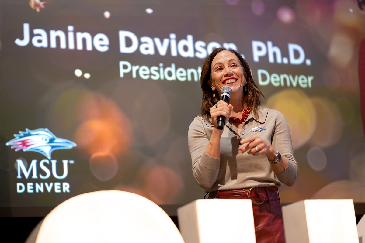 MSU Denver President Janine Davidson speaks into a microphone on stage in front of a projected background displaying her name and title.