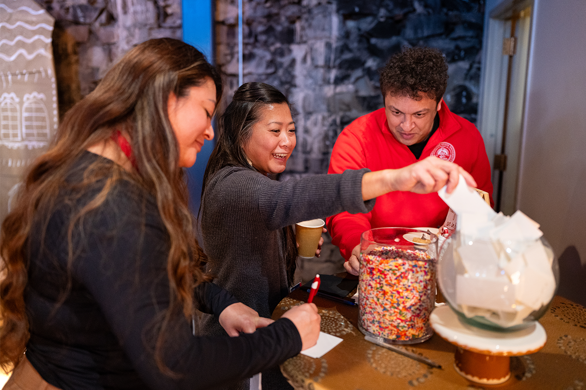 A group of three MSU Denver employees smiles and laughs while writing notes and placing them into a large container filled with colorful candy sprinkles during a Holiday Fest activity.