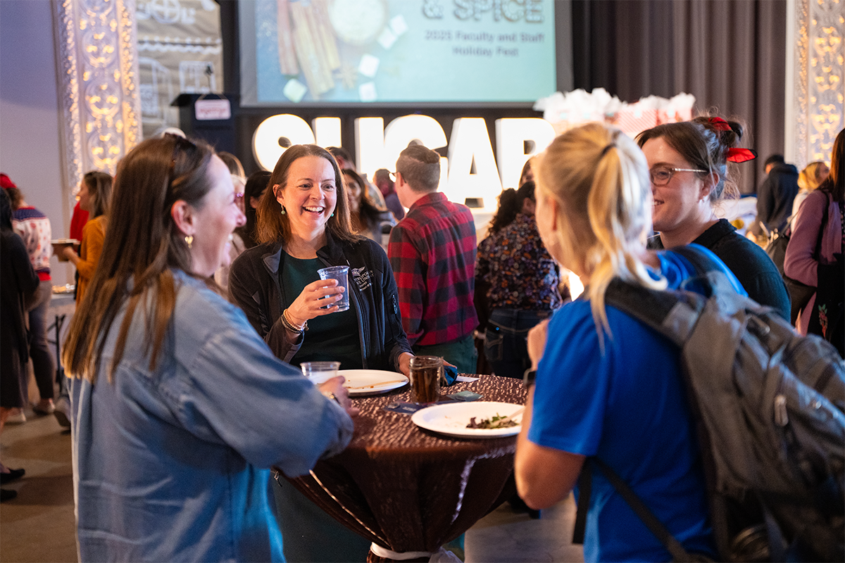 People gathered around a high-top table at an MSU Denver holiday event, smiling and chatting with food and drinks, with festive decor and a large “SUGAR” sign visible in the background.
