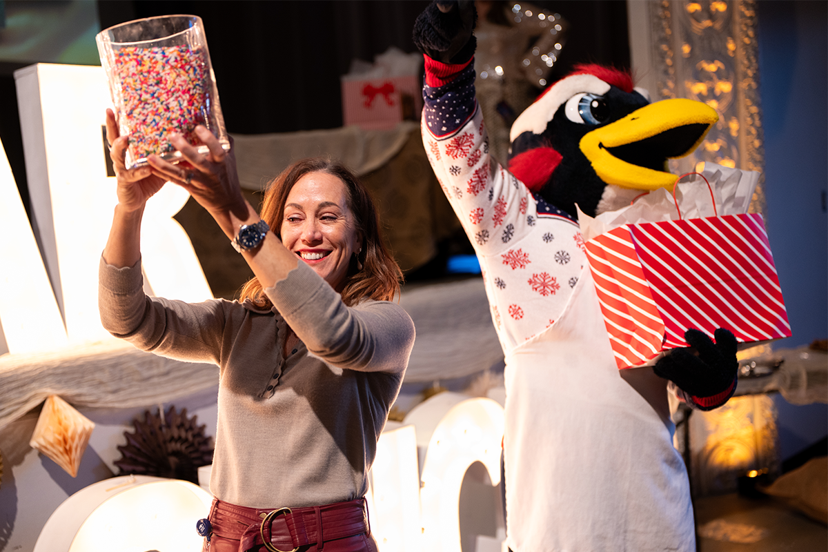 MSU Denver President Janine Davidson stands on stage holding up a large container of colorful sprinkles while the Rowdy mascot beside her lifts a red-and-white gift box during a Holiday Fest celebration.