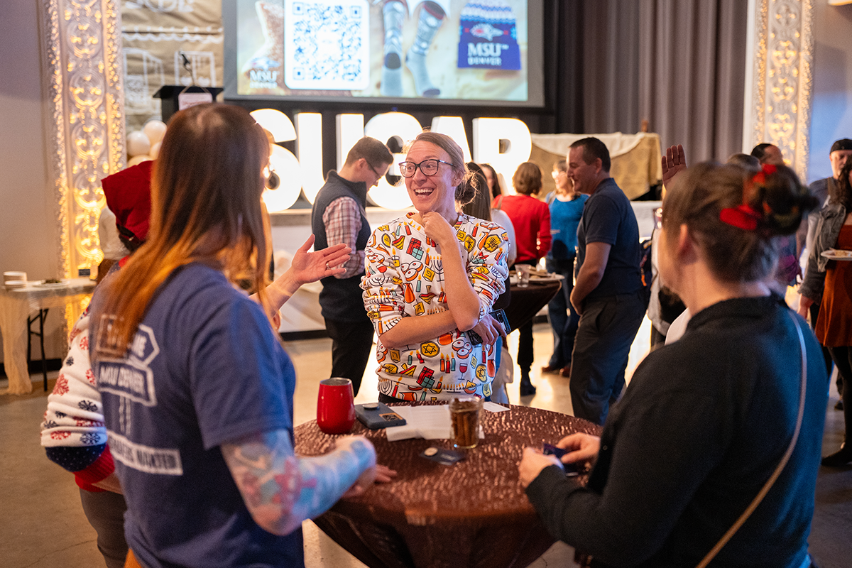 A group of MSU Denver employees stands around a tall table at the Holiday Fest, laughing and talking as they enjoy the decorated event space.