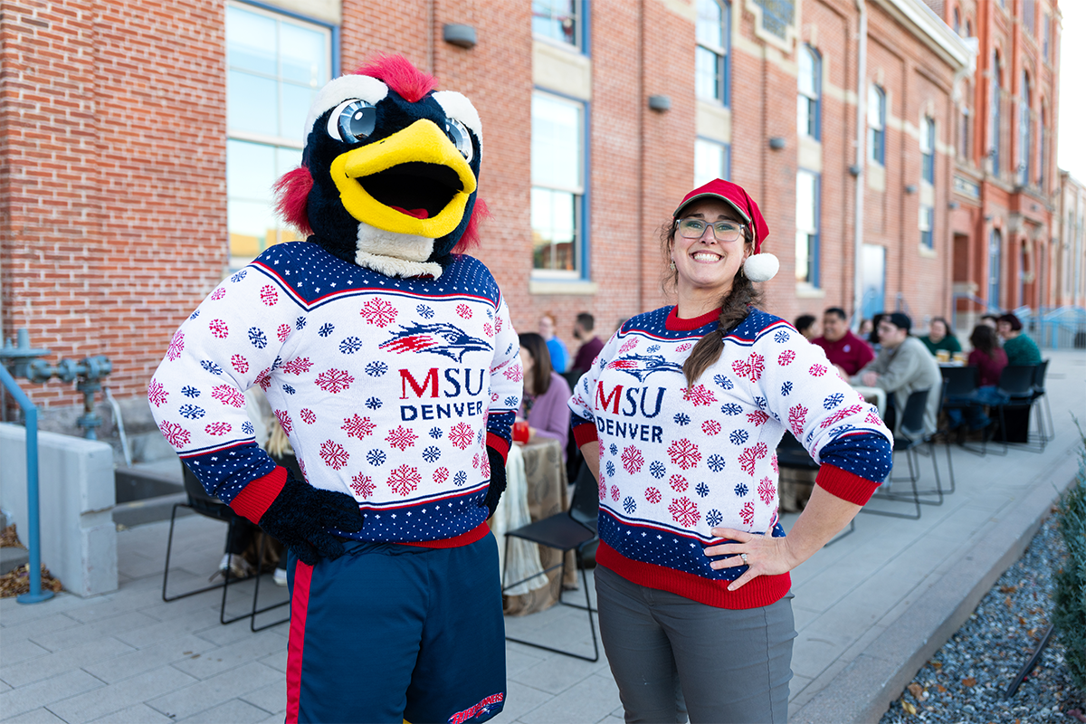 Two people wearing matching MSU Denver holiday sweaters, one in a Rowdy the Roadrunner mascot costume and one in a Santa hat pose outdoors on campus with hands on hips and people seated at tables in the background.
