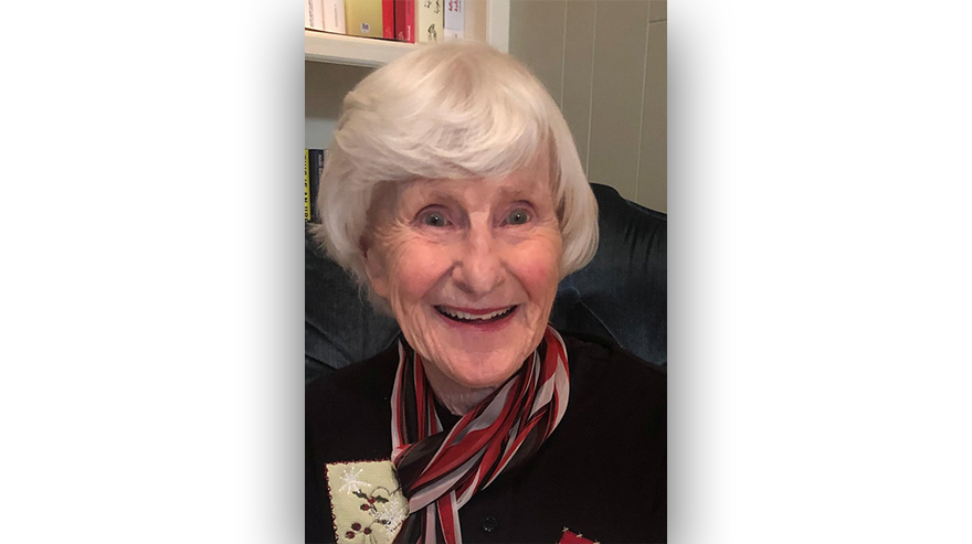 Portrait of Oralie McAffe, smiling warmly while seated indoors, wearing a dark top and striped scarf.