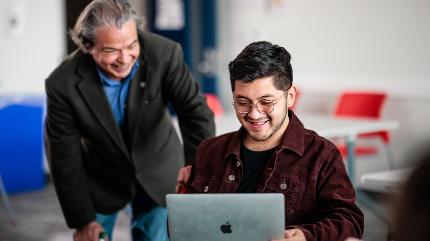 MSU Denver student Gabriel Balbuena Trujillo works on a laptop in a classroom while a professor looks on and smiles.