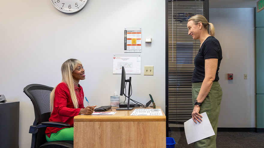 Supervisor and employee talk during a mid-year review in an office, with one person seated at a desk and the other standing and holding papers