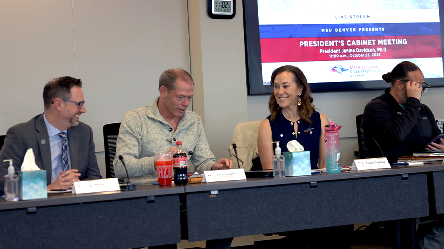 A panel of four MSU Denver leaders sit at a long table during a President’s Cabinet meeting, speaking and taking notes.