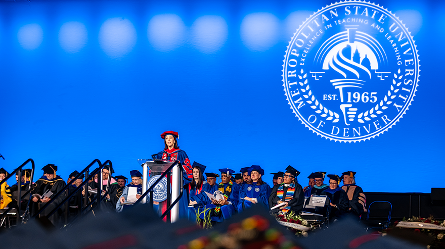 MSU Denver President Janine Davidson delivers a commencement speech onstage in full academic regalia, with faculty and graduates seated behind her in front of a large blue MSU Denver seal.