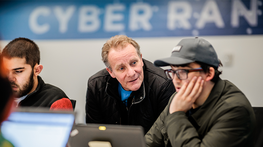 Students working on computers with an instructor providing guidance in a classroom.