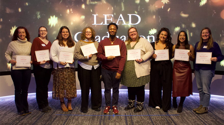 A group of LEAD program graduates stand in a row holding certificates, smiling in front of a backdrop that reads “LEAD Graduation.”