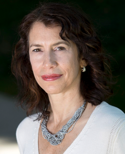 Terry Bower smiling softly in an outdoor headshot, wearing a white top and a silver necklace with a dark green background.