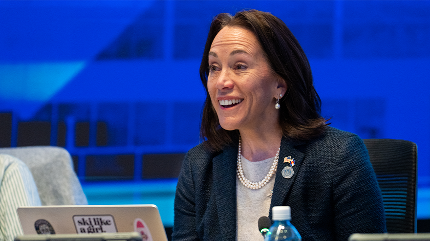 Janine Davidson speaks during a Board of Trustees meeting while seated at a table with a laptop and microphone.