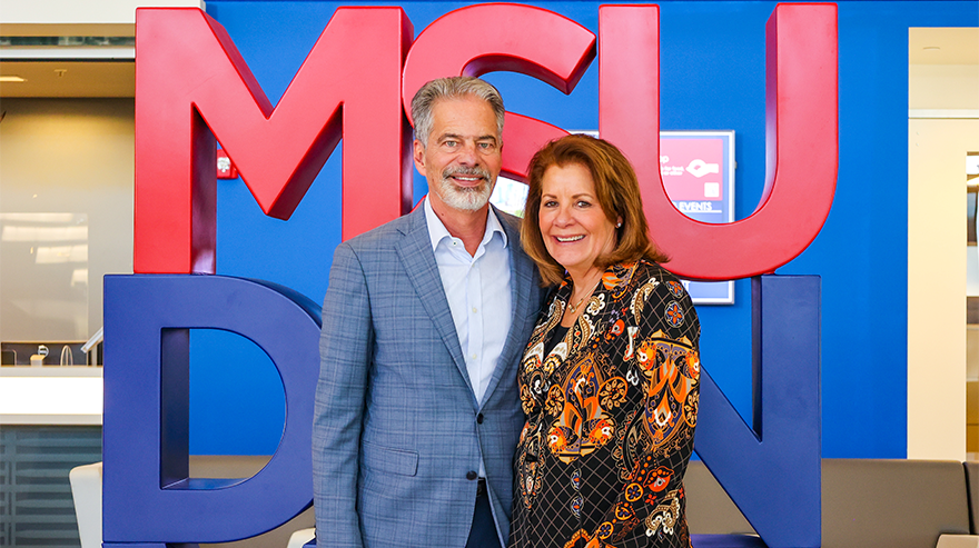 Two donors stand smiling in front of a large MSU Denver sign on campus.