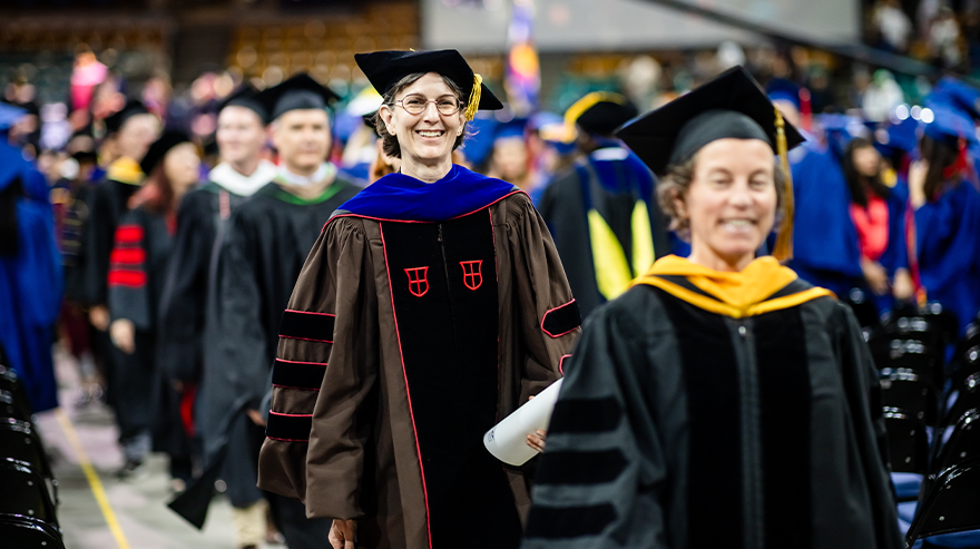Faculty members in academic regalia walk in a procession during an MSU Denver commencement ceremony, surrounded by graduates and attendees in the arena.