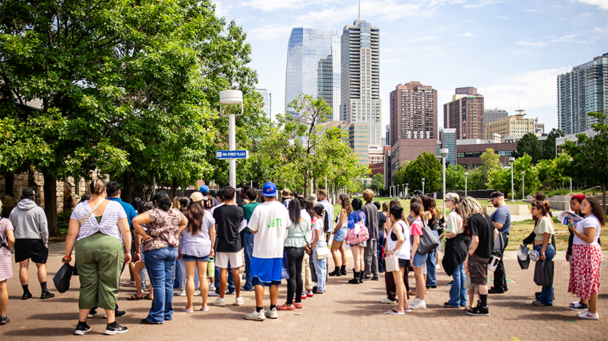 Group of students and visitors gathered on a campus walkway with the Denver skyline visible in the background on a sunny day.