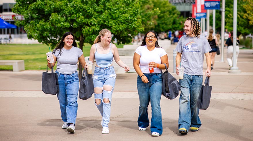 Four students walk together across campus carrying tote bags and drinks, smiling and talking.
