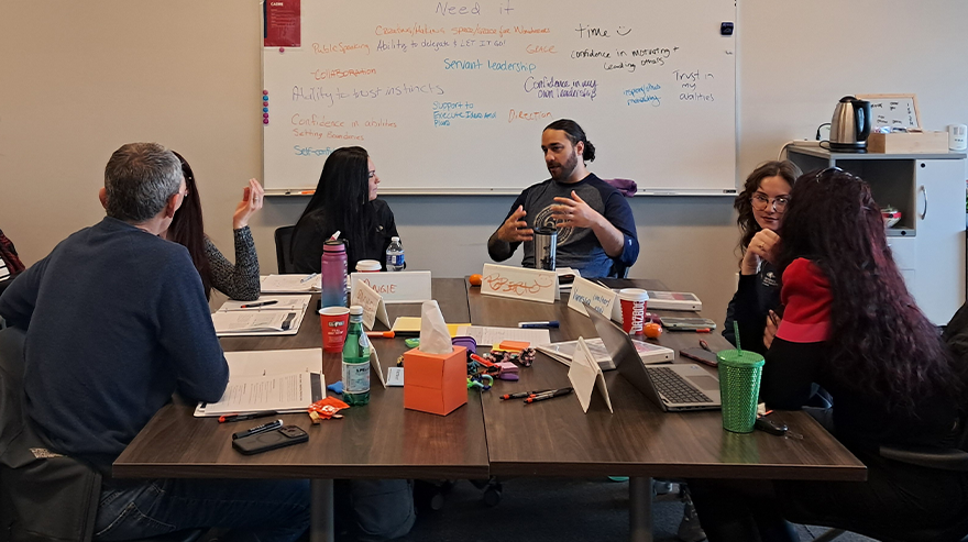 Group of students and instructors sit around a table in a classroom discussion with notebooks, laptops, and drinks visible, while a whiteboard with notes is behind them.