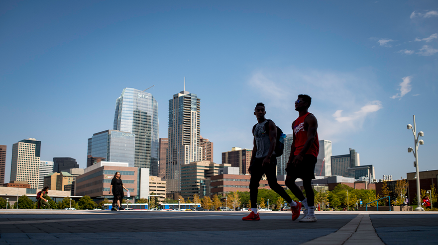 Two students walk across campus with the Denver skyline visible in the background on a clear day.