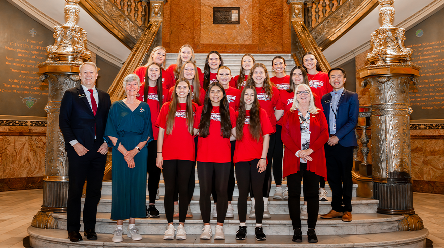 Group of students in matching red shirts and staff members pose on a staircase inside a government building with ornate columns and architecture.