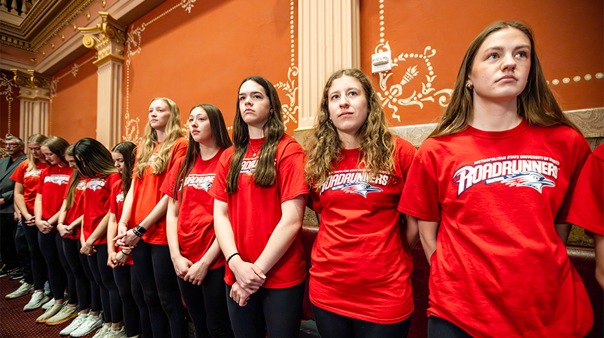 Students wearing red Roadrunners shirts stand in a line inside a government building, listening attentively during an event.