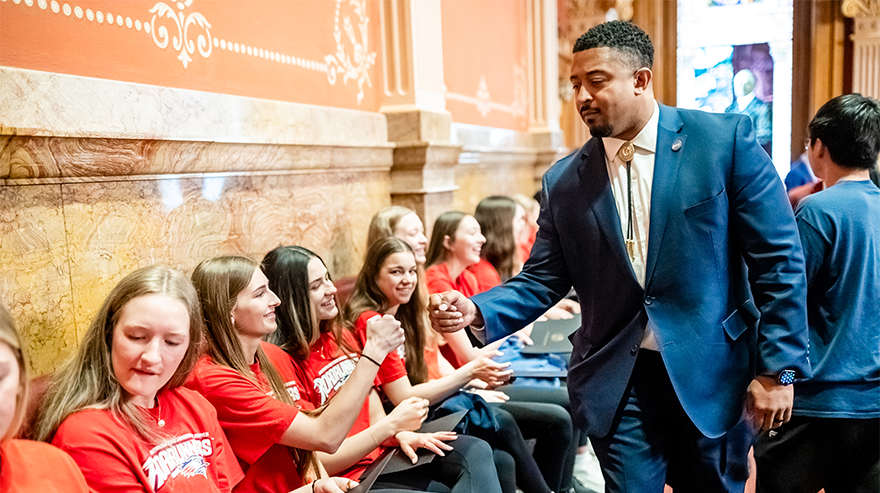 Man in a suit greets and fist-bumps students wearing red Roadrunners shirts while they sit in a government building during an event.