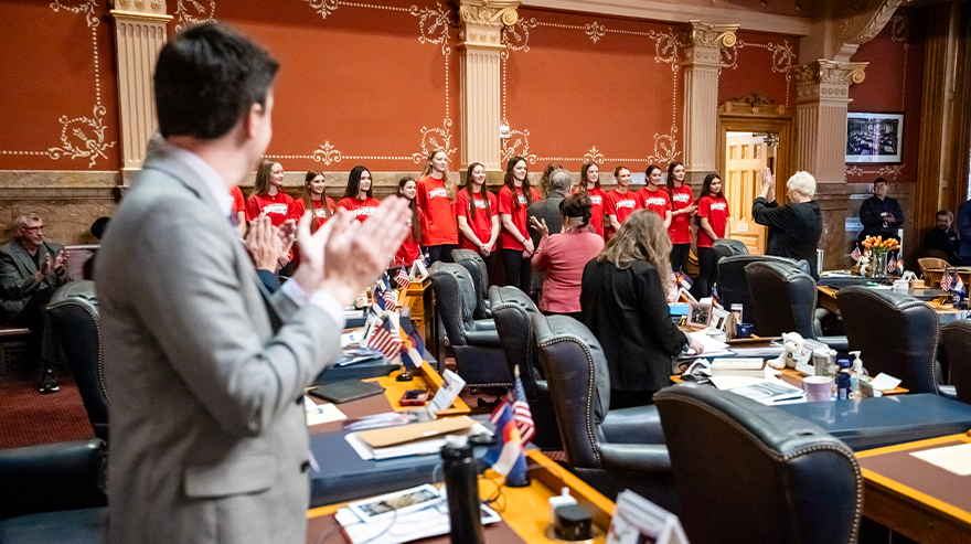 Students wearing red Roadrunners shirts stand in a line inside a government building, listening attentively during an event.