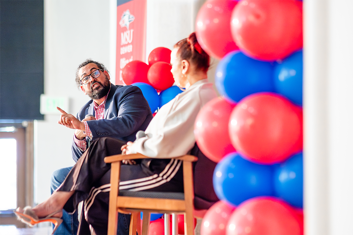 Two speakers sit on stage in conversation during an MSU Denver event, with red and blue balloons in the background.