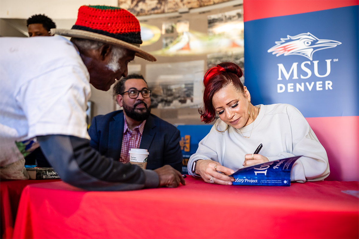 Woman signs a book at a table during an event while two people stand nearby, with an MSU Denver banner in the background.