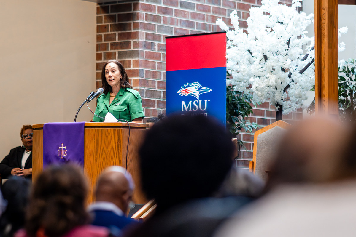 Speaker stands at a podium addressing an audience during the Rachel B. Noel community event.