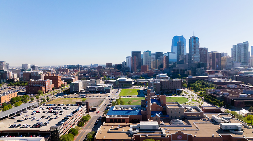 Aerial view of the Auraria Campus and downtown Denver skyline on a clear day, showing academic buildings, green spaces, parking areas, and surrounding city high-rises.