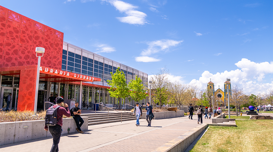 Students walk and gather outside the Auraria Library on the Auraria Campus on a sunny day with trees and campus buildings in the background.