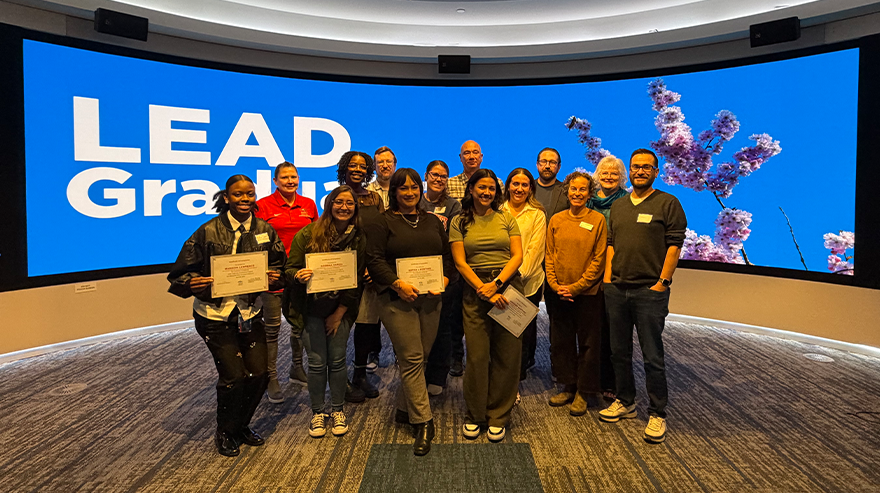 Group of students and staff posing with certificates in front of a large screen reading “LEAD Grad” during a graduation celebration event.