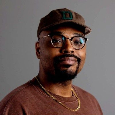 Headshot of Branden Whitfield wearing a brown hat, glasses, a brown shirt and a gold necklace