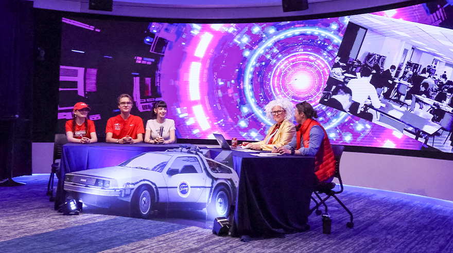Panel of speakers seated at tables in a studio space with a colorful digital backdrop and a display car prop, presenting during a live event or broadcast.