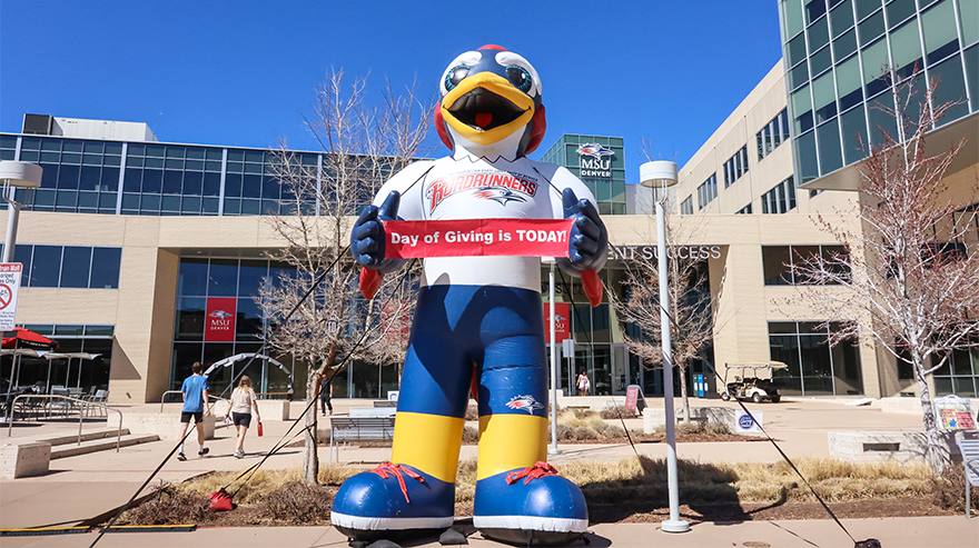 MSU Denver Roadrunner mascot holding a “Day of Giving is TODAY!” sign outside a campus building on a sunny day.