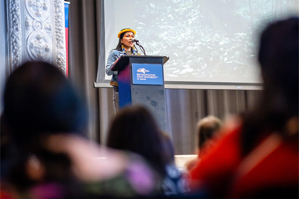 Speaker stands at a podium addressing an audience, with the crowd blurred in the foreground and a presentation screen behind her.